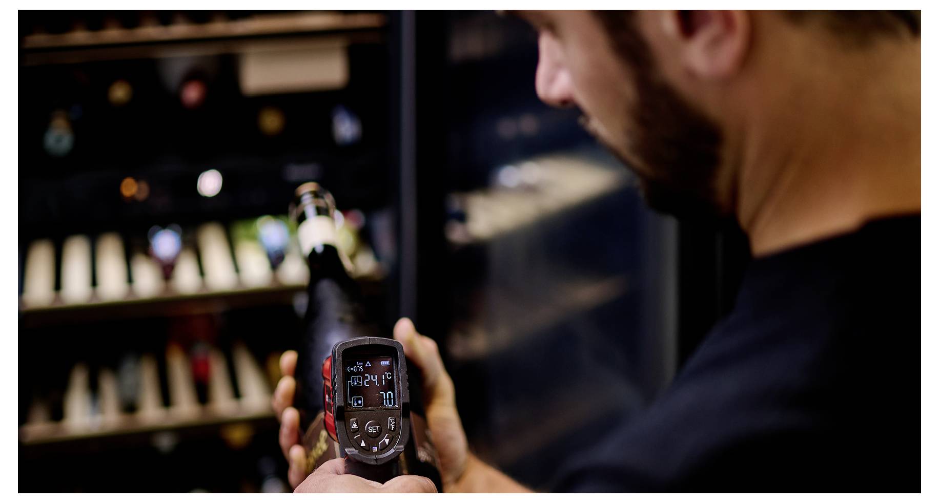 A person holds a wine bottle with a digital thermometer attached, showing 12°C. Background shows a wine fridge with bottles.