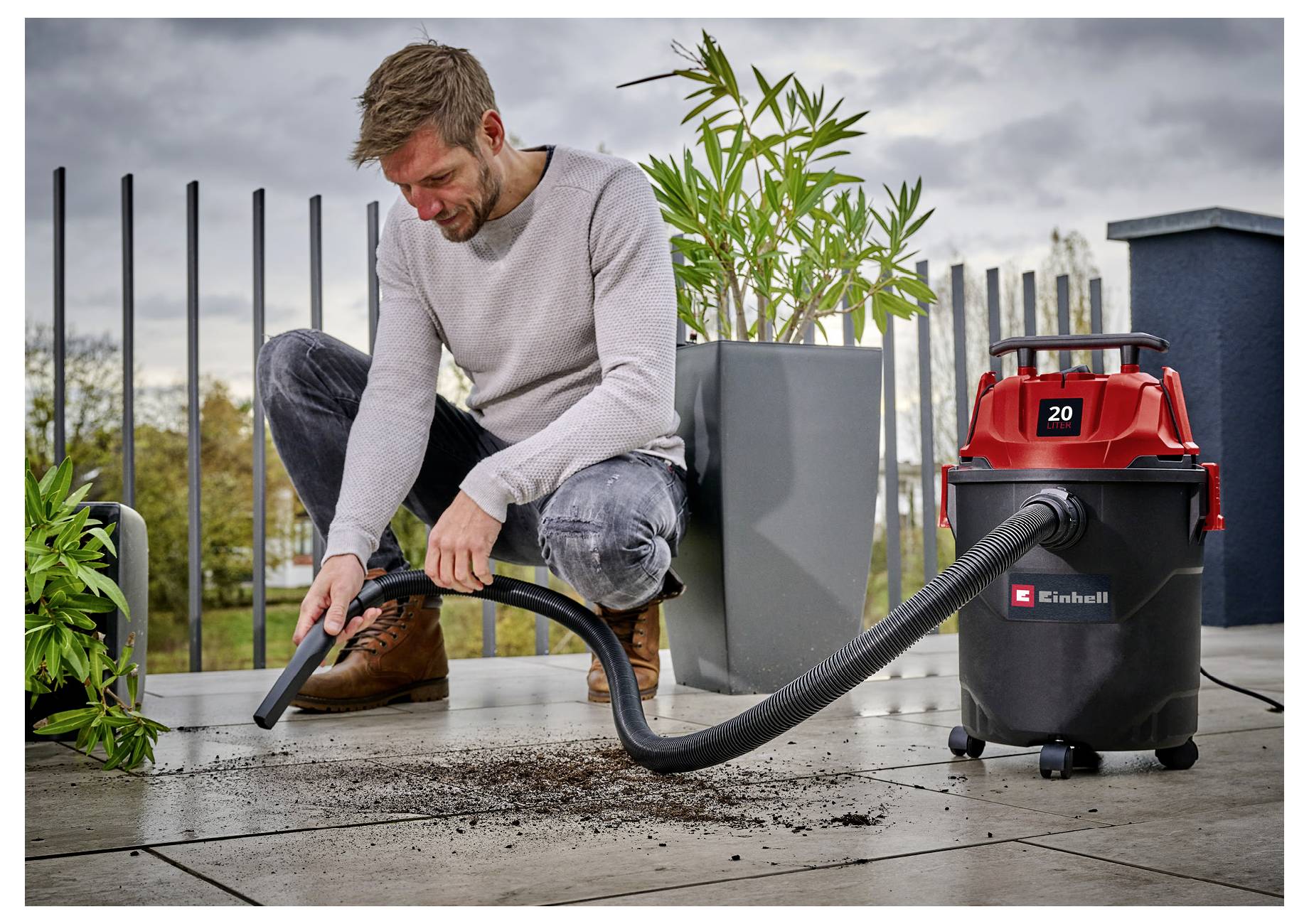 A man kneeling on a terrace uses a red and black vacuum cleaner to clean up spilled soil next to a potted plant on a cloudy day.
