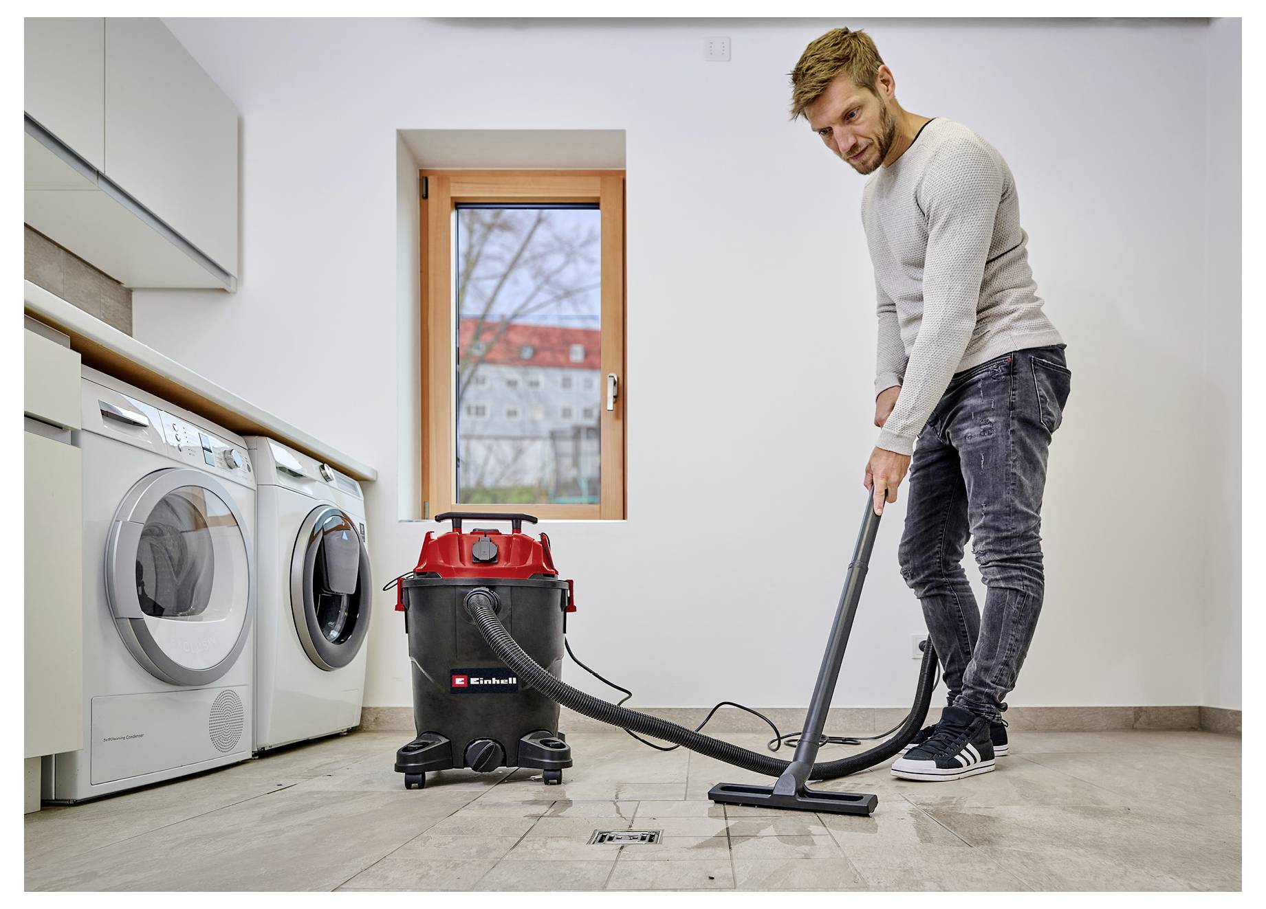 A person vacuuming a laundry room floor with a red and black vacuum cleaner, standing near a washing machine and dryer by a window.