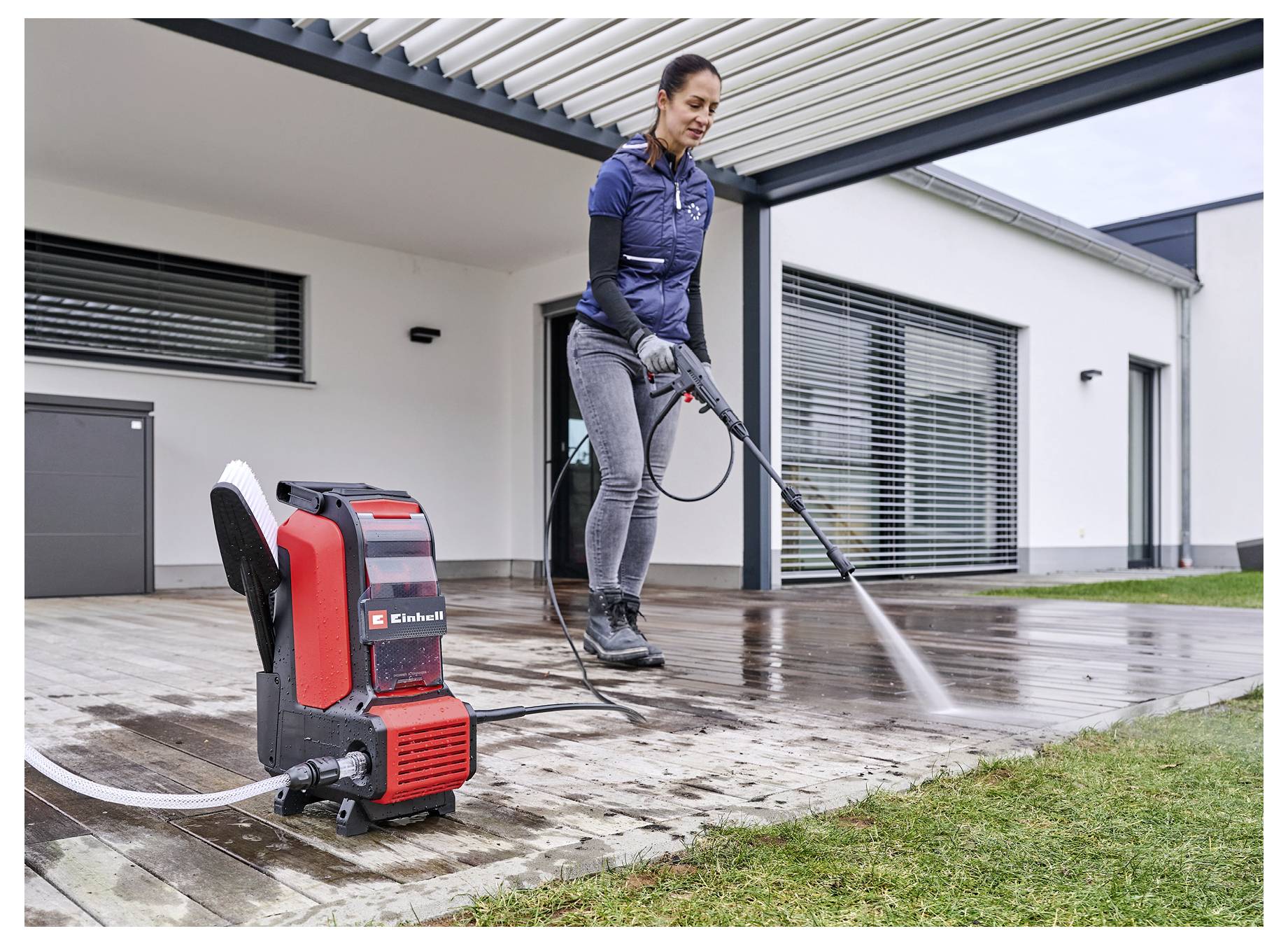 A person is using a power washer to clean a wooden deck attached to a modern white house. The washer is branded and red.