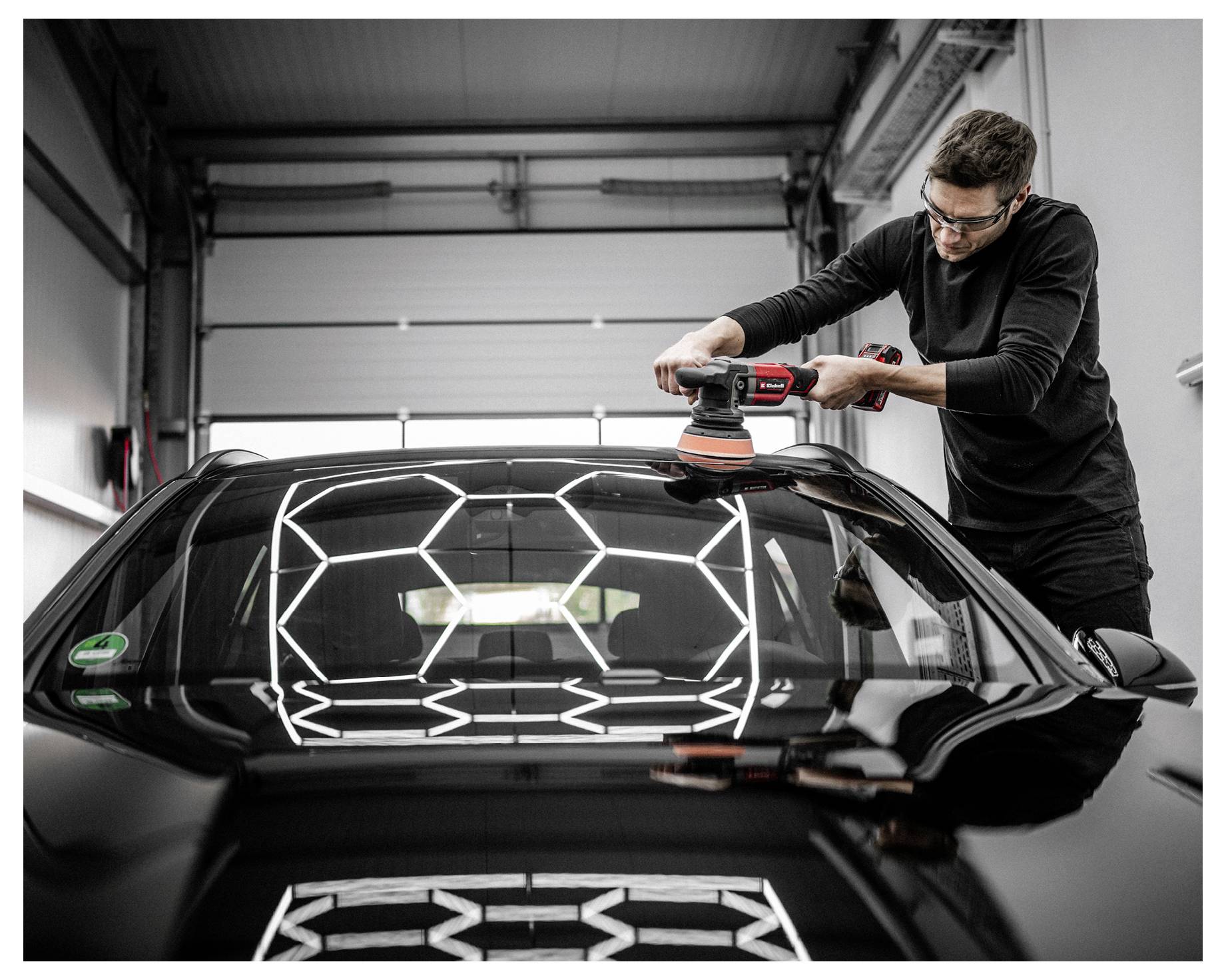 A person polishes the roof of a black car inside a garage, reflecting bright hexagonal lights, emphasizing a shiny finish.