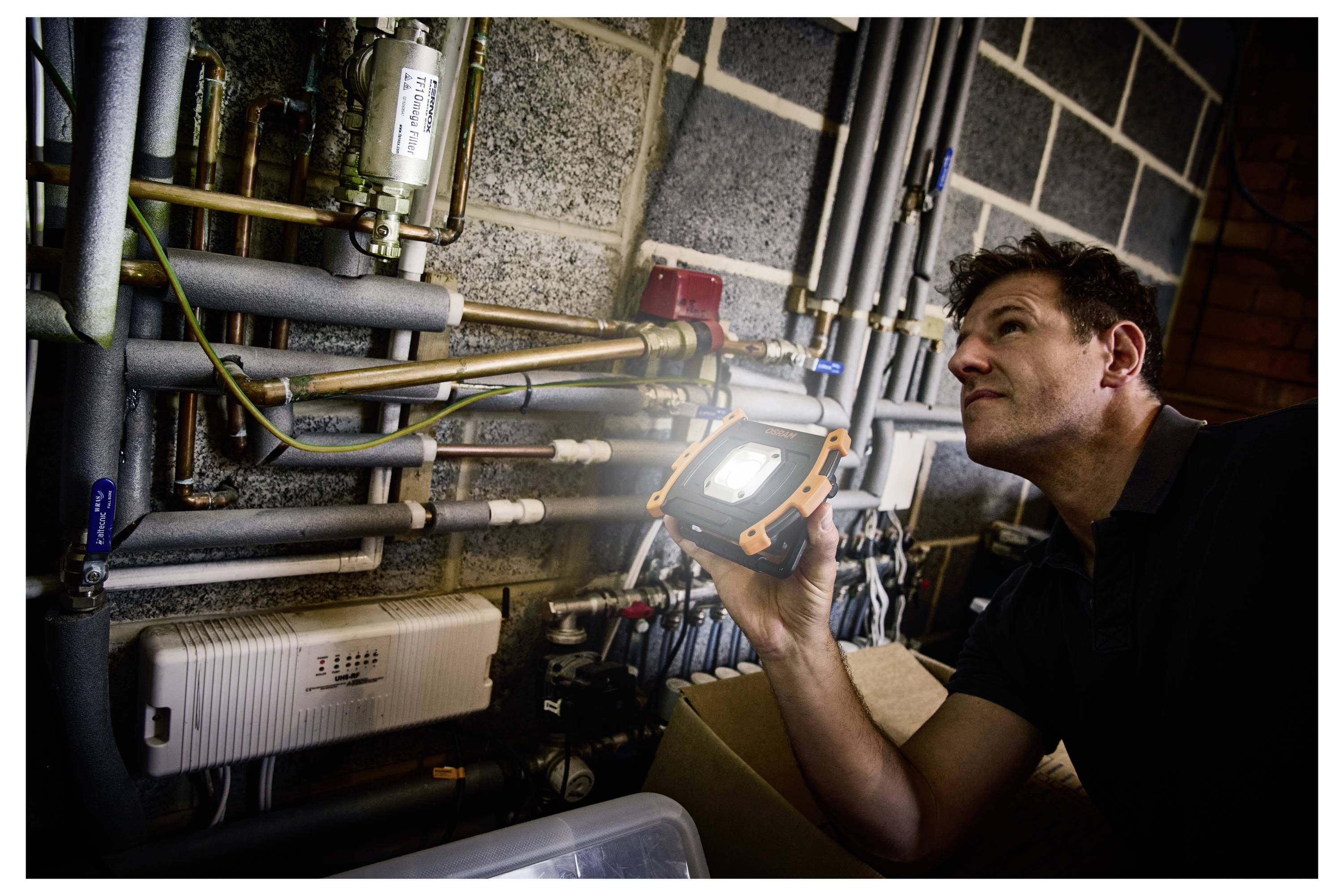 A person inspects complex plumbing and heating pipes in a dim room using a portable light, focused intently on the system.