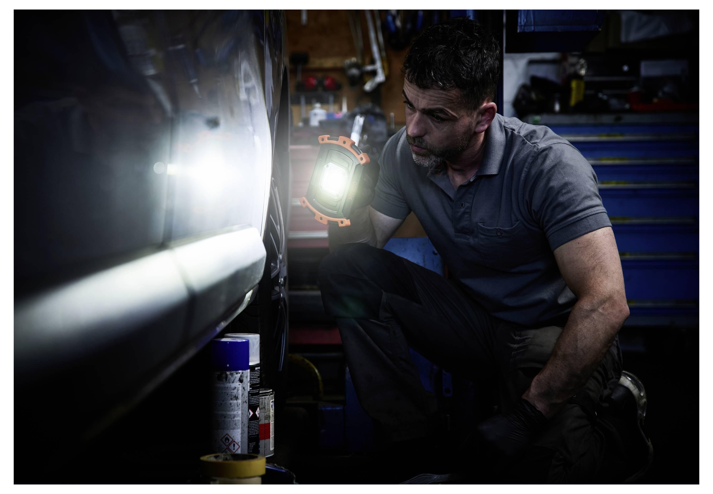 A mechanic crouches next to a vehicle, using a bright flashlight to inspect its side, surrounded by tools in a workshop setting.