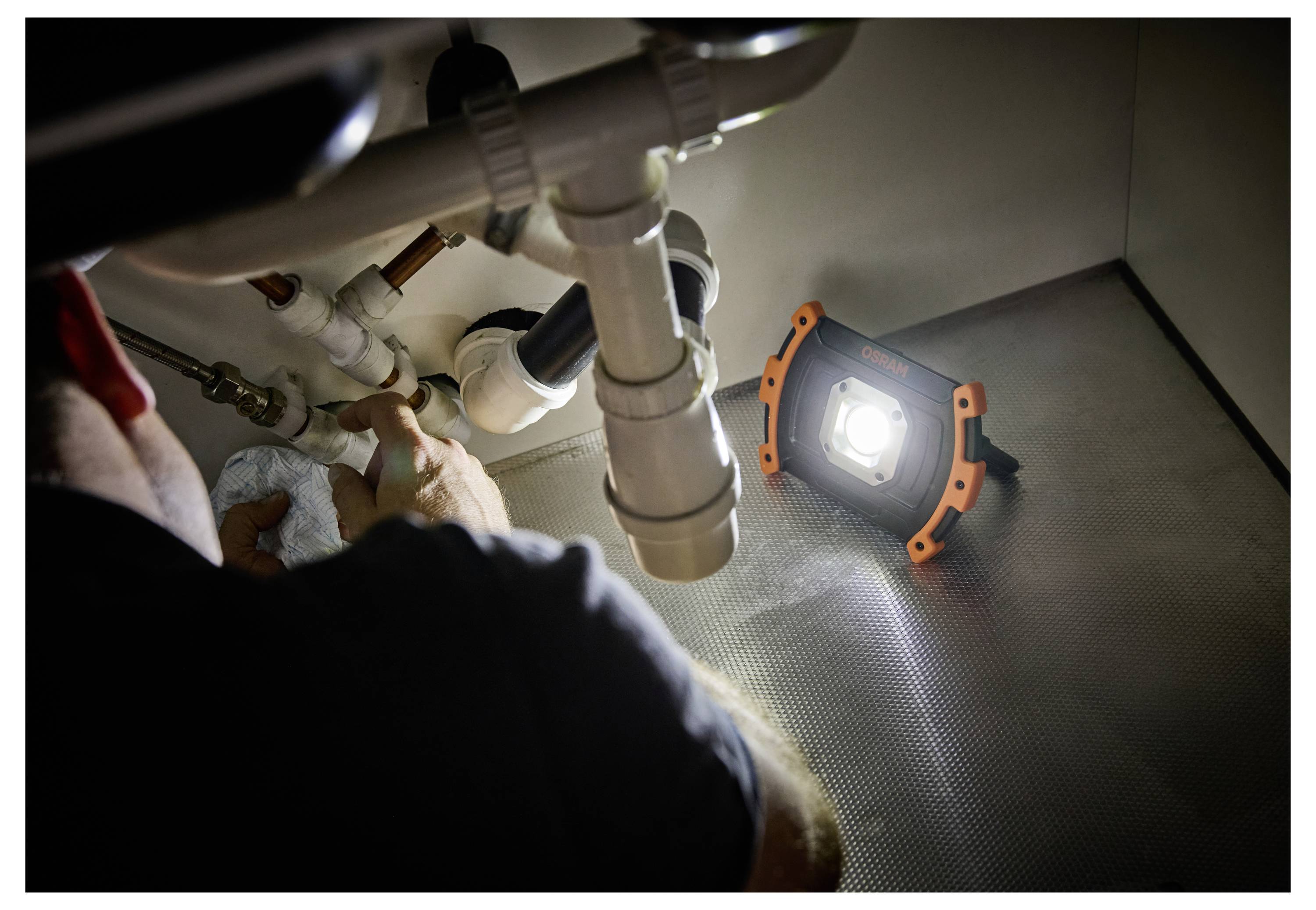 A person repairs plumbing under a sink, illuminated by a portable work light placed on the floor, highlighting pipes and tools.