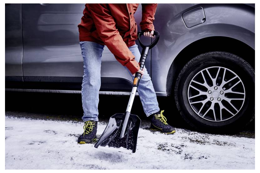 A person wearing a red jacket and jeans uses a shovel to clear snow from a driveway next to a silver car, focusing on winter cleanup.