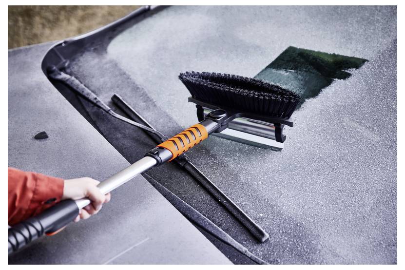 A hand uses a squeegee with a brush to clean frost from a car windshield, revealing a clear patch amidst the icy surface.