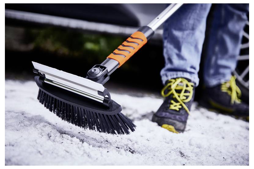 A person brushes snow from the ground using a tool with a brush and squeegee attachment. The squeegee is orange, and the person wears yellow-laced shoes.