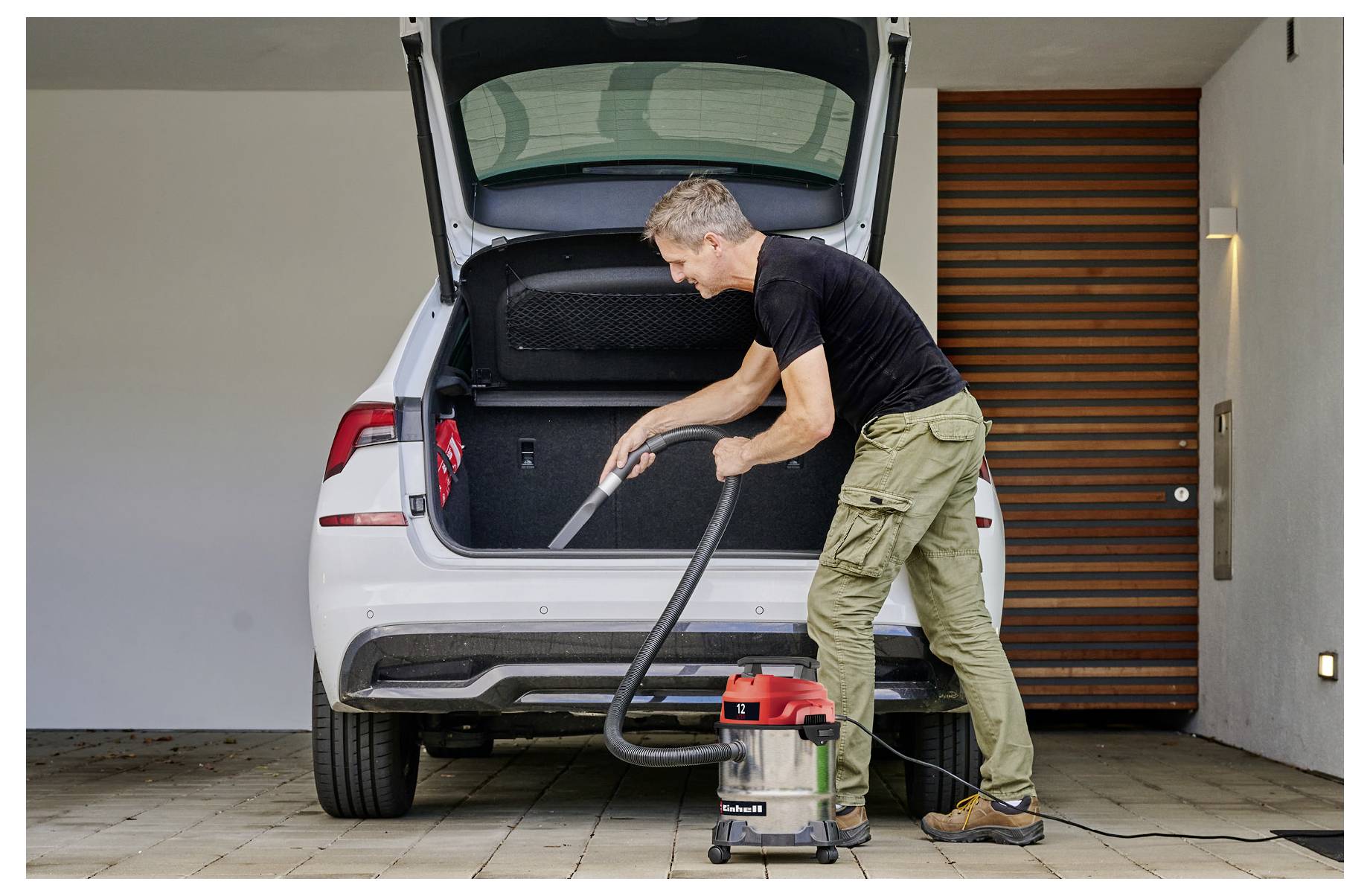 A person vacuums the trunk of a car in a garage, using a red and grey vacuum cleaner. The car's hatchback is open, revealing a black interior.