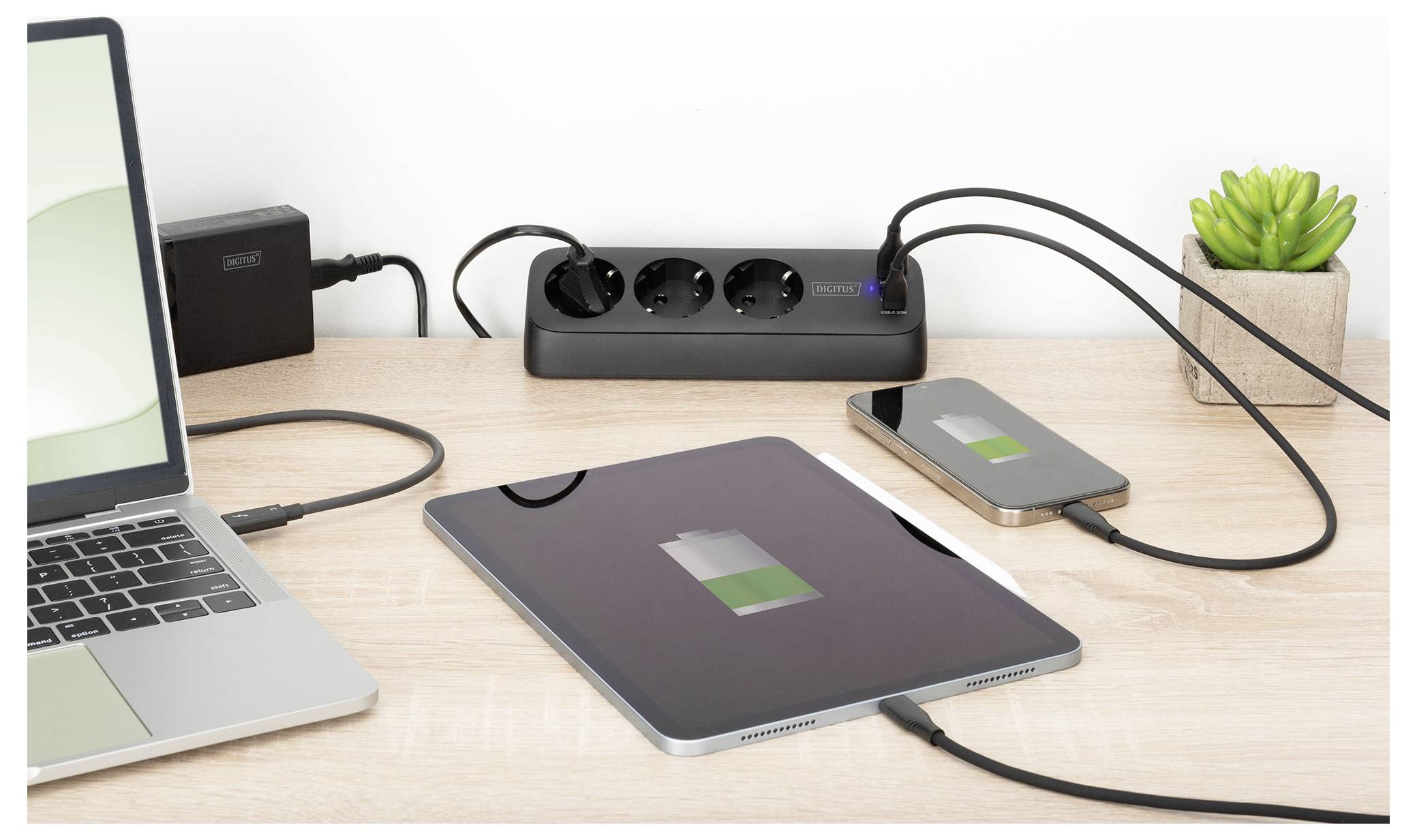 A tablet and smartphone charging on a desk, connected to a power strip alongside a laptop. A small potted plant is in the background.