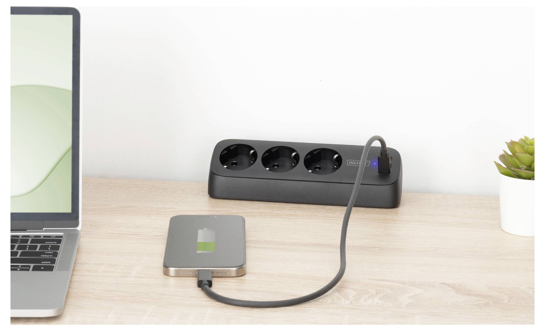 A laptop on a desk is connected by a cable to an external device showing a charging symbol. A power strip with three outlets is in the background.