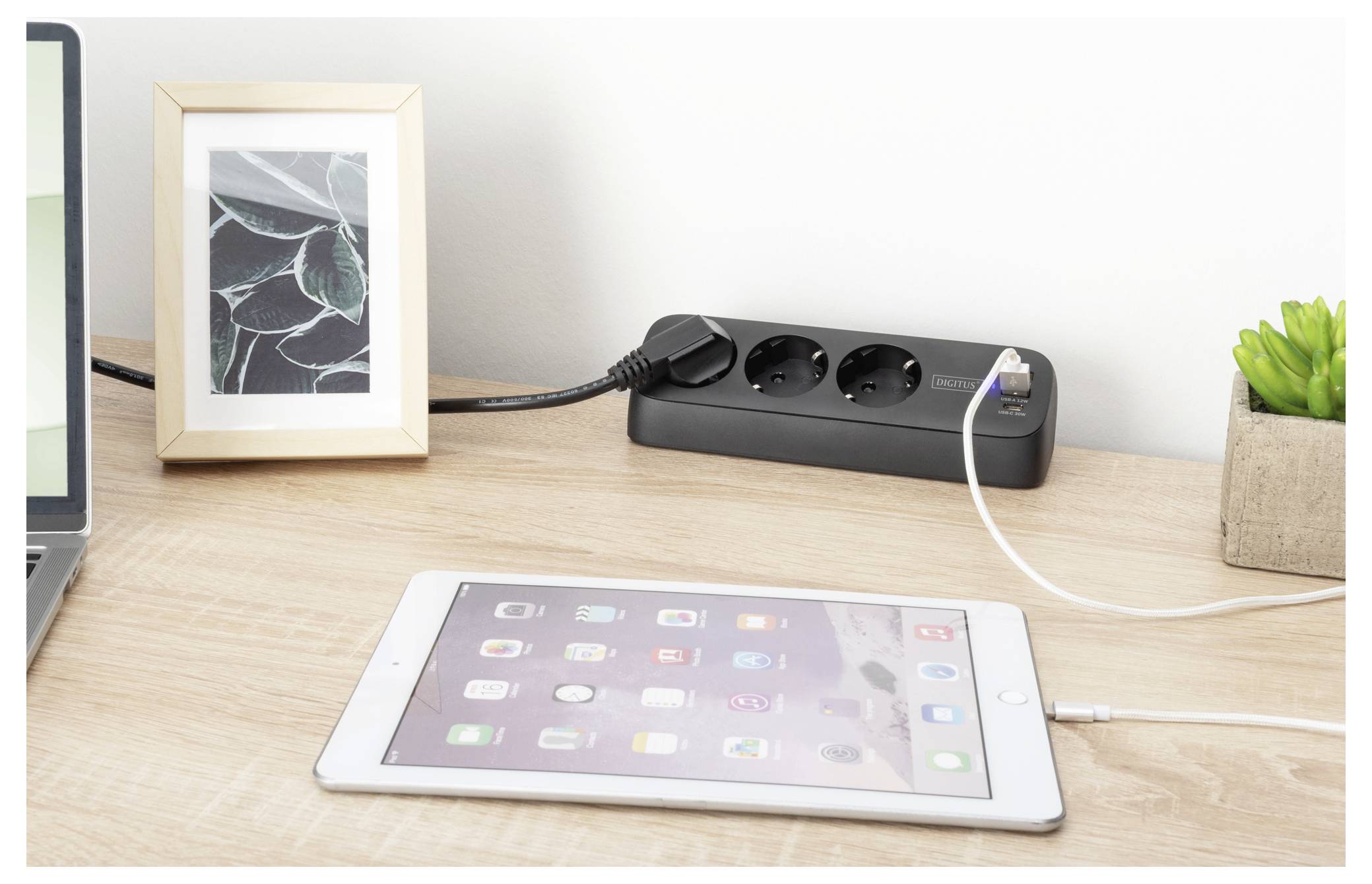 A white tablet charging on a wooden desk beside a framed art piece, succulent plant, and power strip with plugged-in devices.