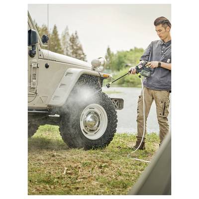 Man washing a beige Jeep with a pressure washer outdoors, spraying the front wheel and fender near a lake.