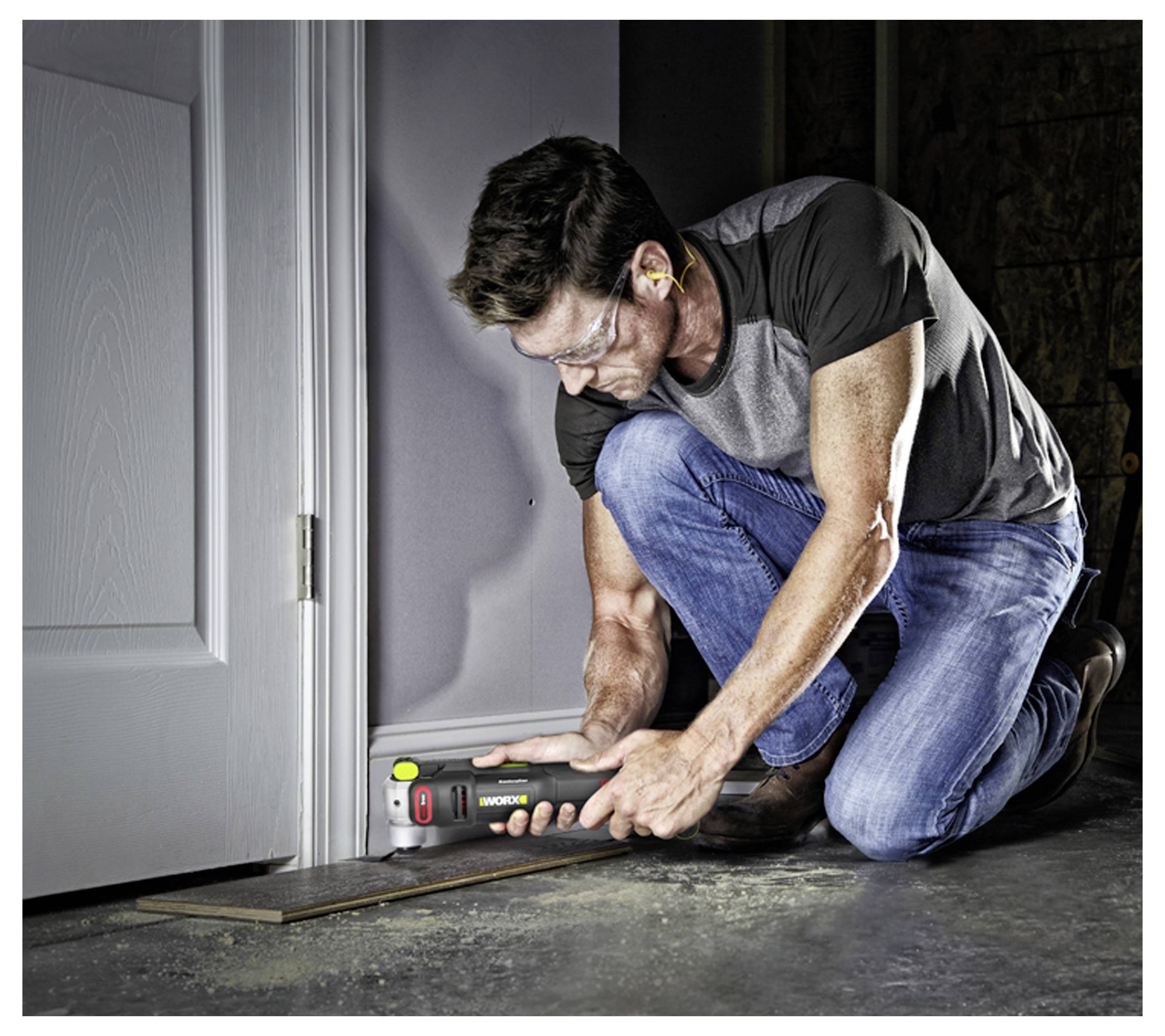 A man kneels on a floor using a power tool to cut or trim near a doorway inside a dim room.