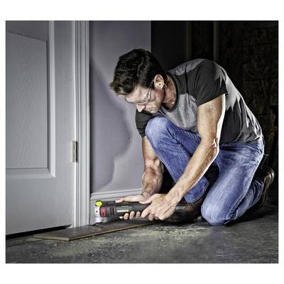 A man kneels on a floor using a power tool to cut or trim near a doorway inside a dim room.