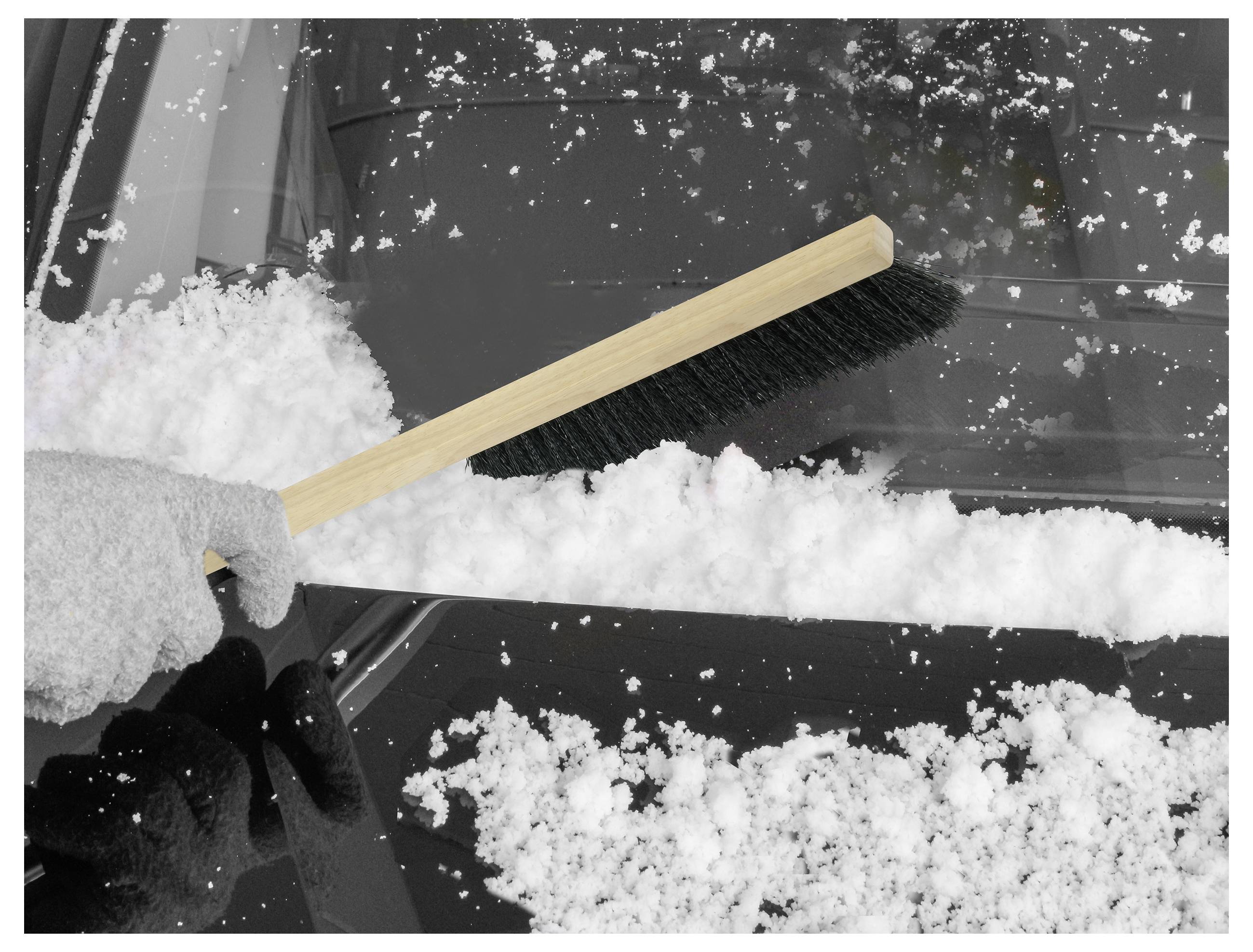 A gloved hand uses a wooden brush to clear snow off a car windshield. The car is covered in a layer of snow, indicating cold weather.