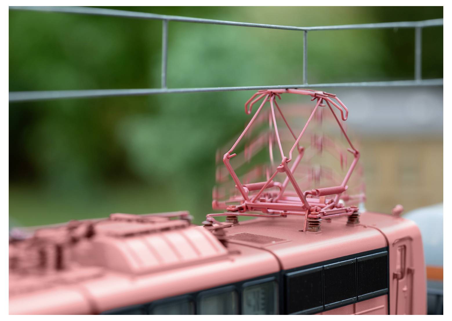 A close-up of a model train's roof, featuring a red pantograph against a blurred green background.