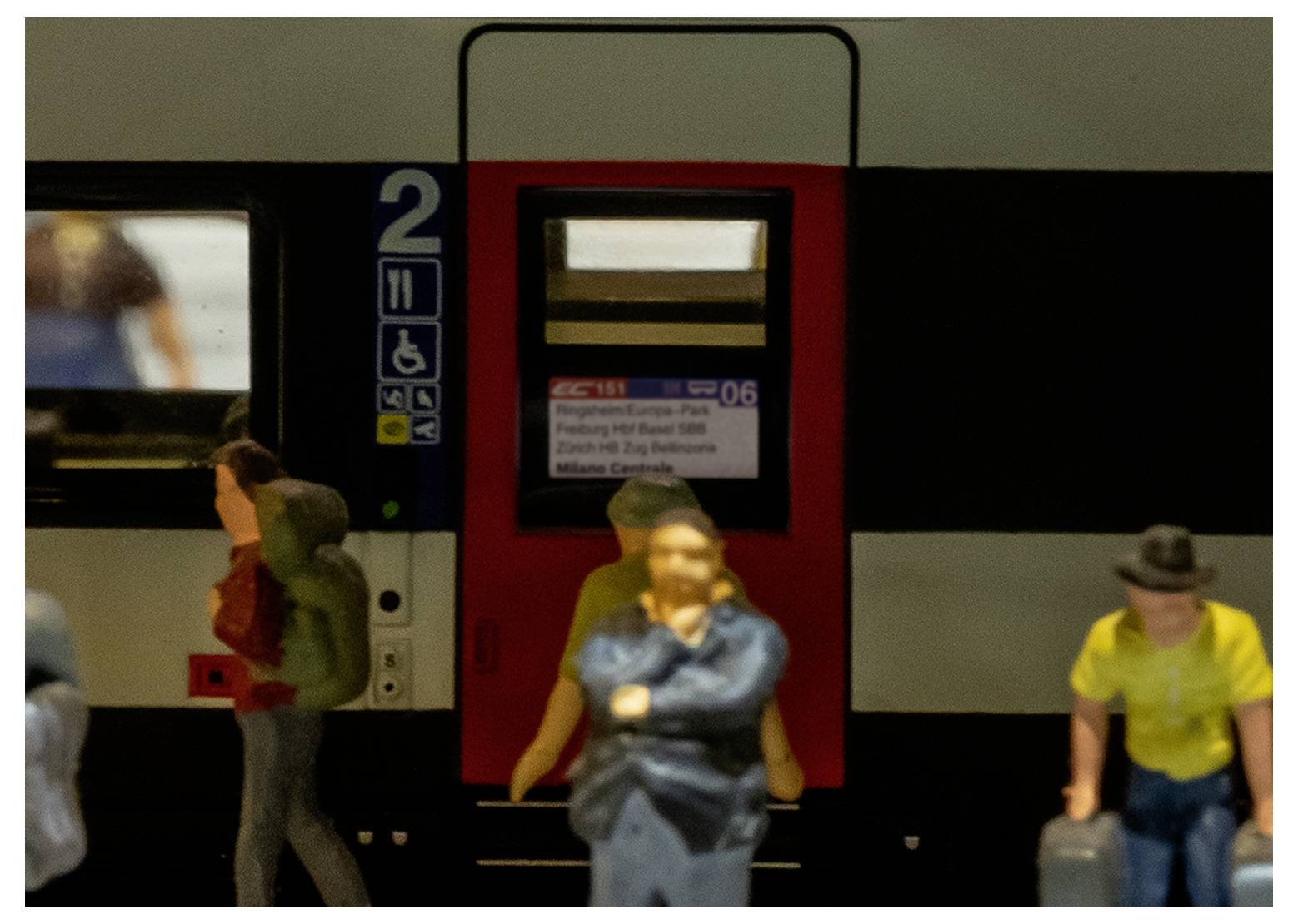A model train with figures stands in front of a train car door. The car number '2' and various symbols indicate accessibility features.