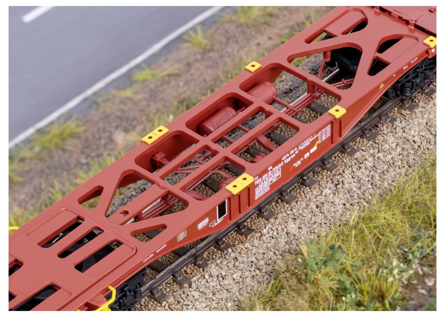 A red, flatbed train car with an open-frame design travels on a track next to a road. It's surrounded by gravel and grass.