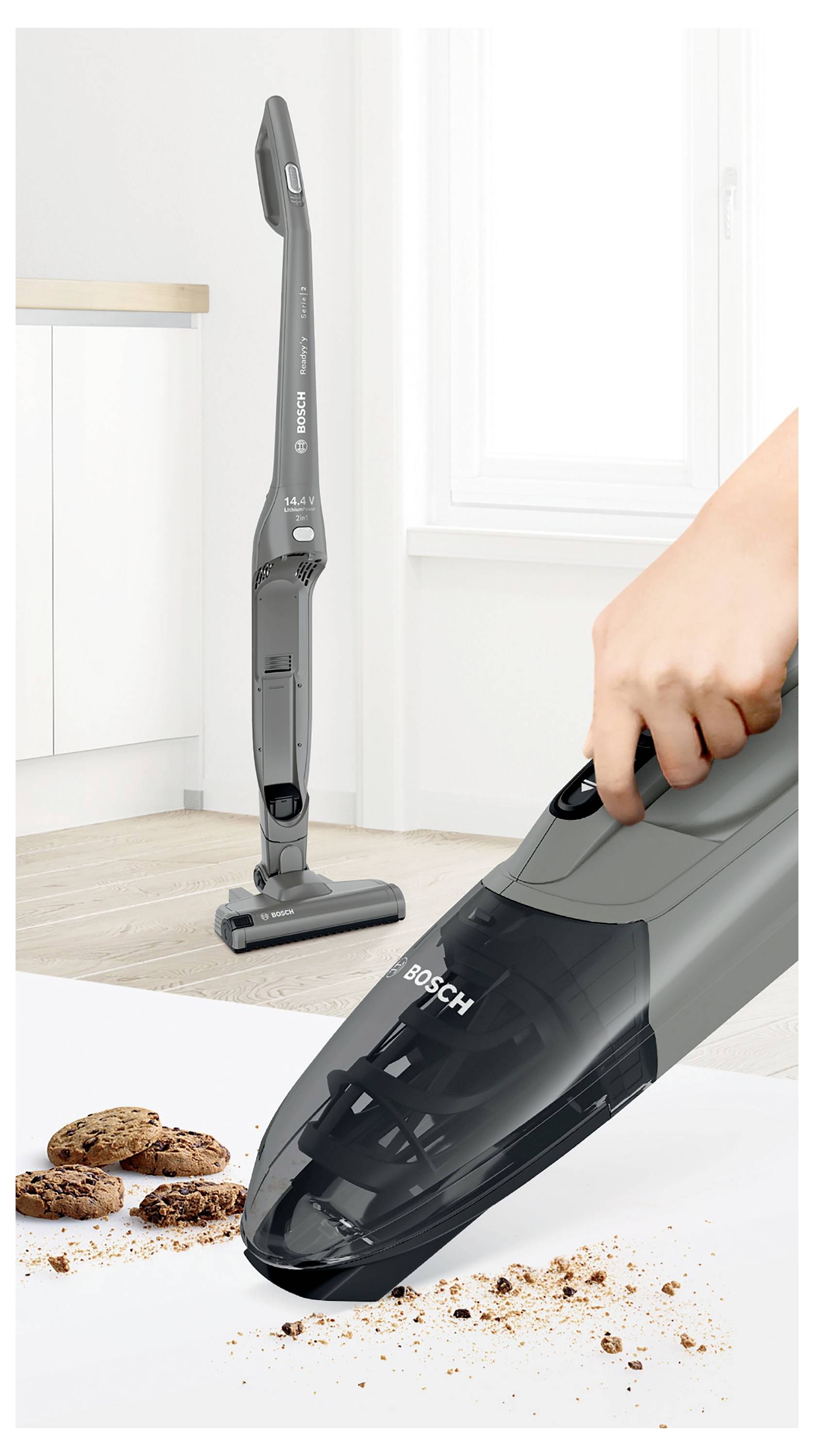 A hand uses a Bosch vacuum cleaner to pick up cookie crumbs from a white surface. Another vacuum stands upright in the background.
