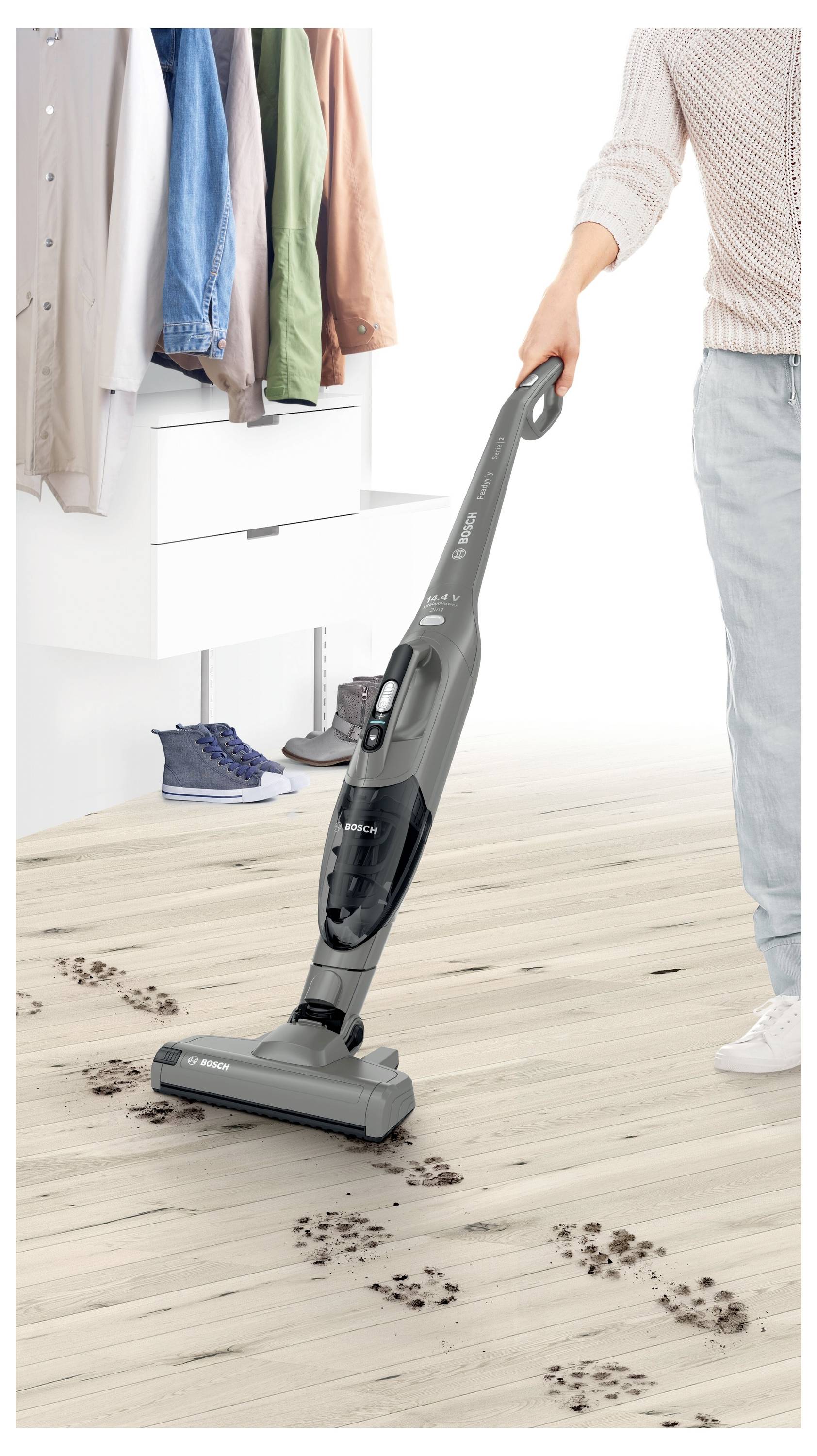 A person uses a cordless vacuum to clean muddy paw prints from a light wood floor in a closet area with hanging clothes and shoes.