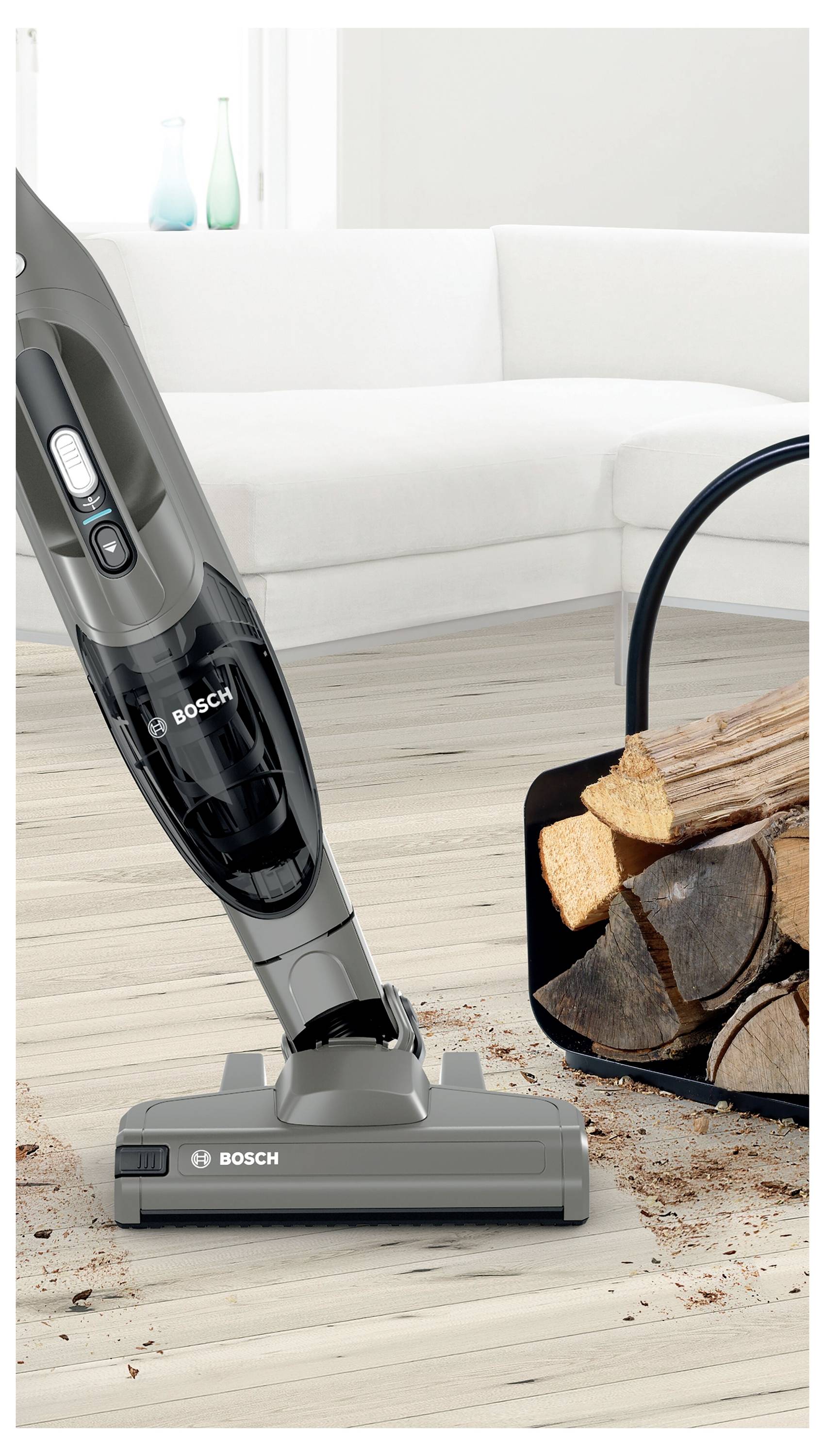 A cordless vacuum cleaner standing upright on a wooden floor next to a basket of firewood in a modern living room setting.