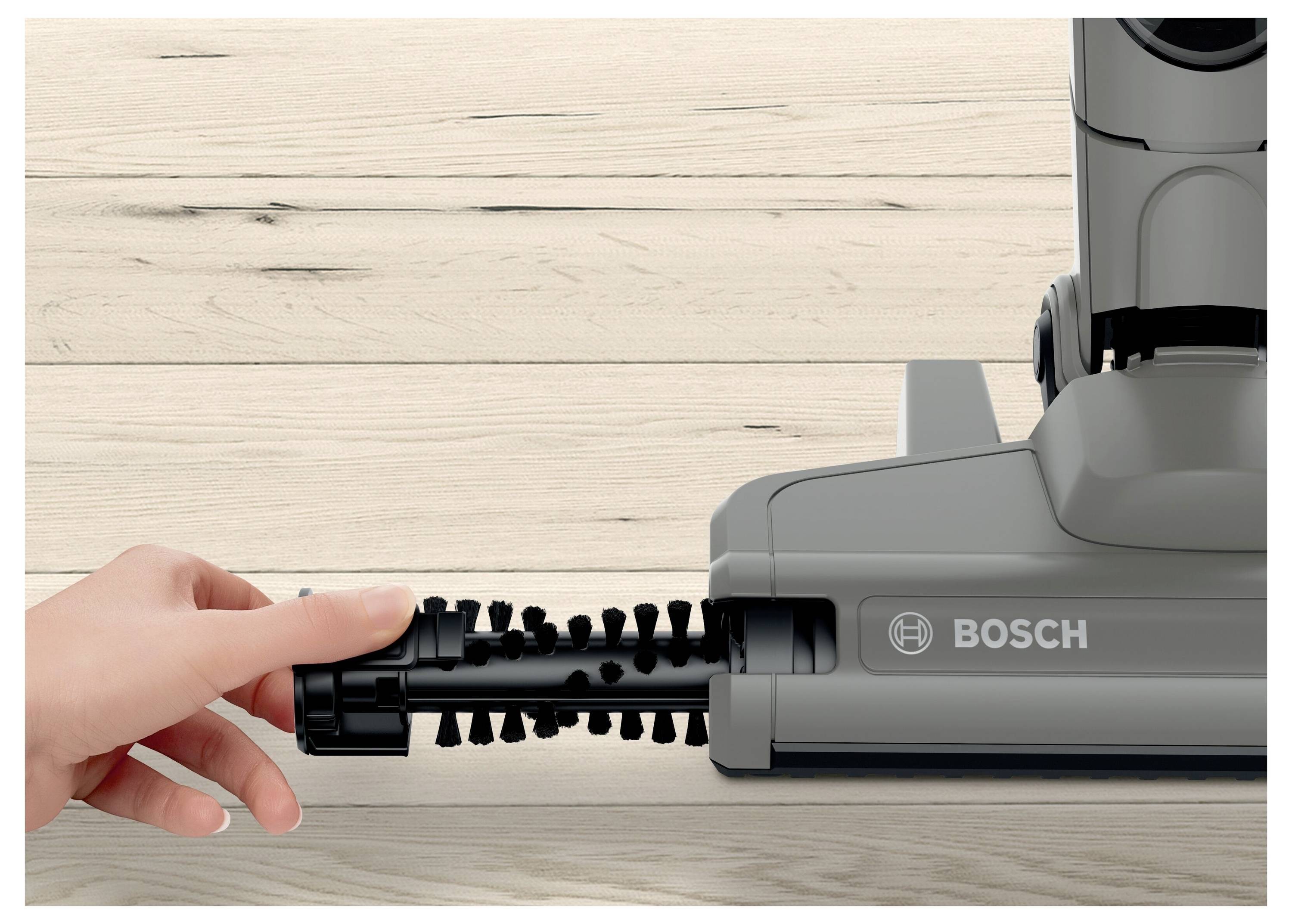 A person pulls a brush from a floor vacuum cleaner on a wood floor. The vacuum has the brand label 'Bosch'.