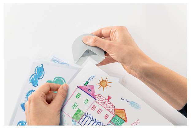 Two hands laminating a colorful drawing of a house using a handheld laminator, with additional drawings underneath.
