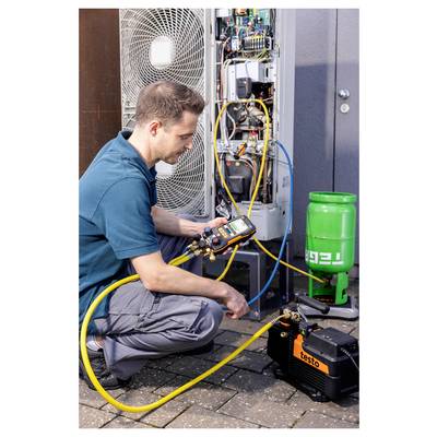 A technician kneels, using a digital manifold to check an HVAC system, with refrigerant gas cylinder and compressor on the ground.