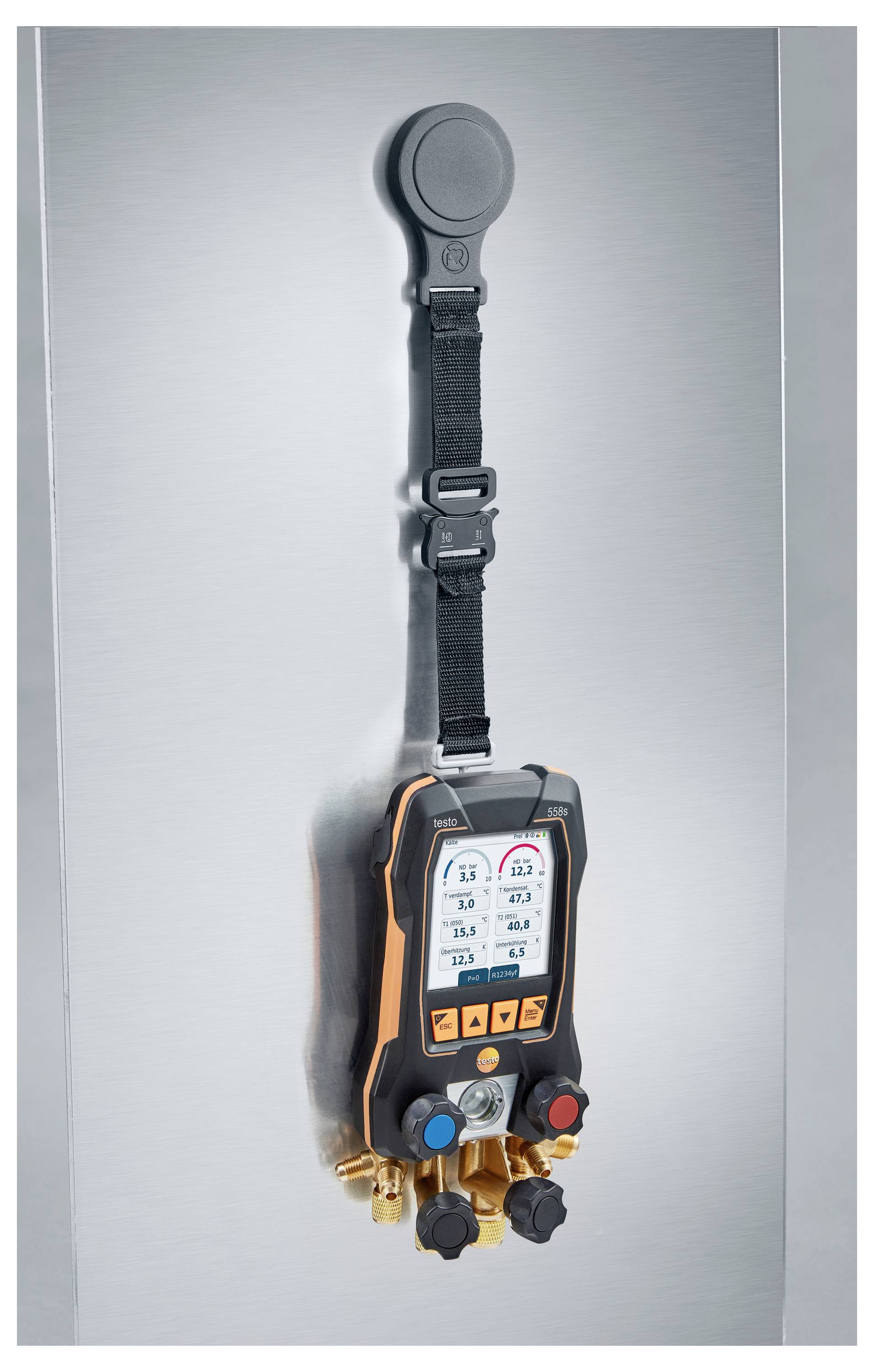 A digital manifold gauge with a display showing measurements, hanging vertically with a strap on a metallic surface.
