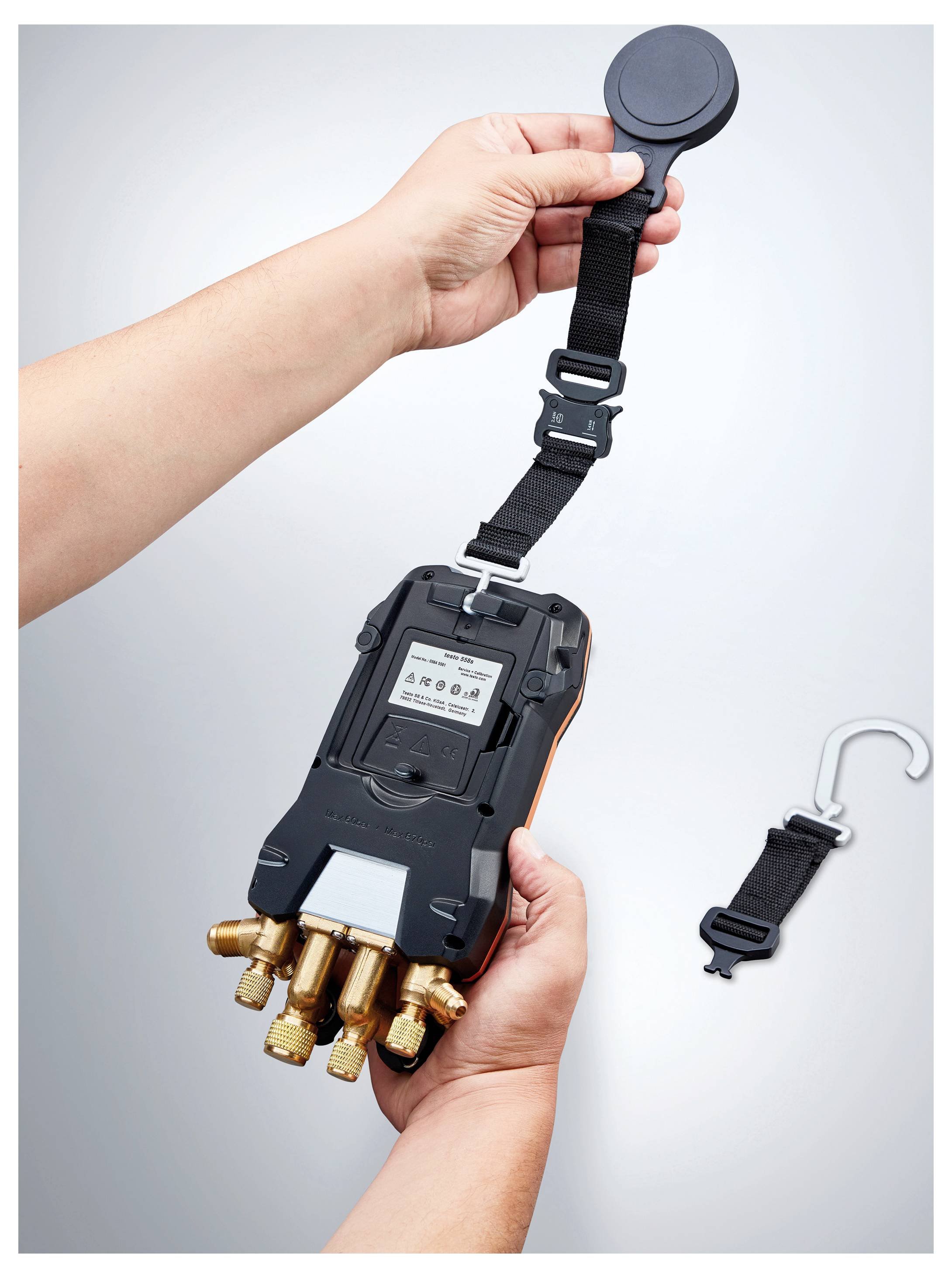 A person holds a handheld device with several brass fittings, attaching a strap with a circular end. A separate hook is placed beside it.