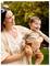 A woman smiling while holding a laughing baby outdoors in a park setting with trees in the background.