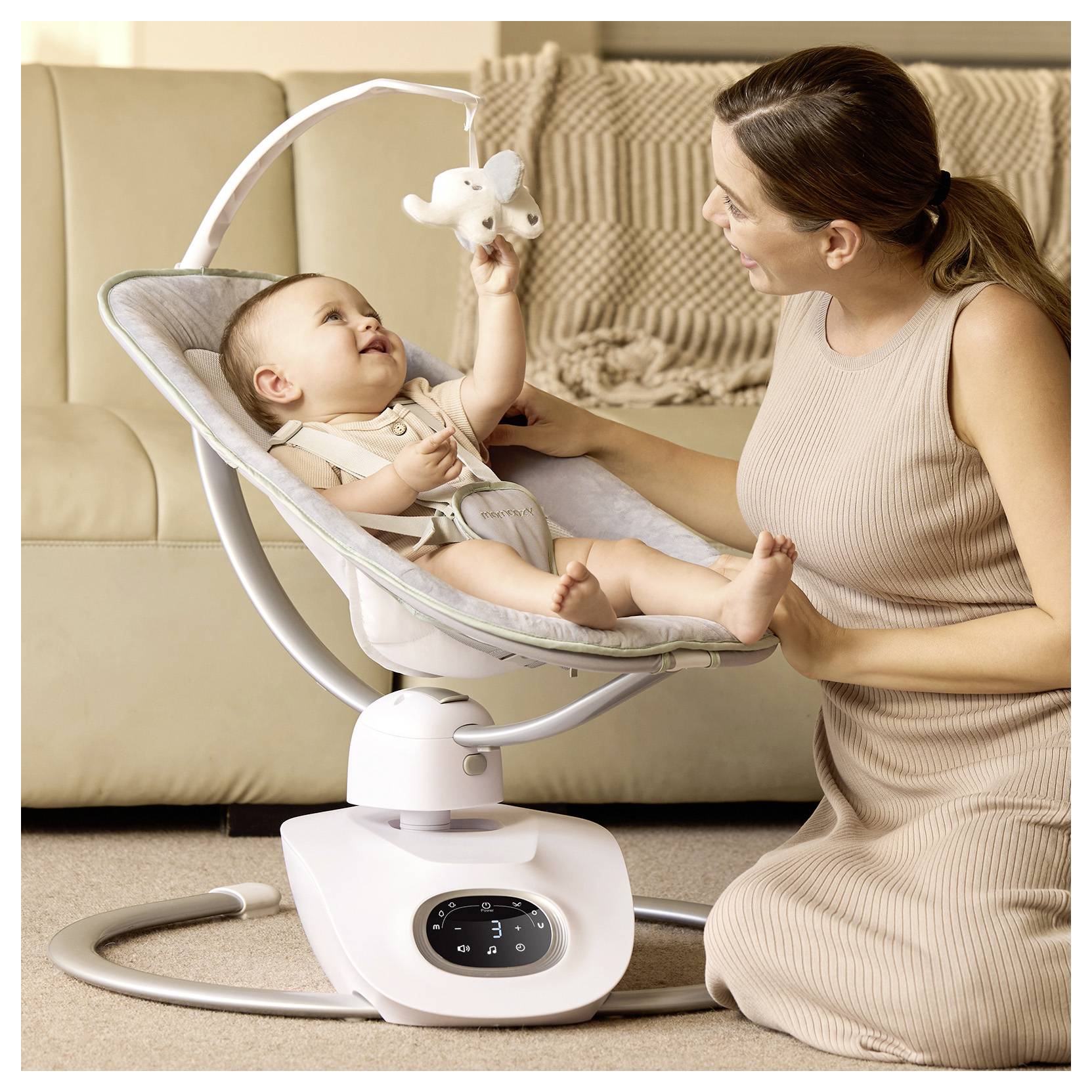 A mother smiles at her baby, who is seated in a baby swing, reaching for a plush toy. They are indoors, creating a warm and nurturing environment.