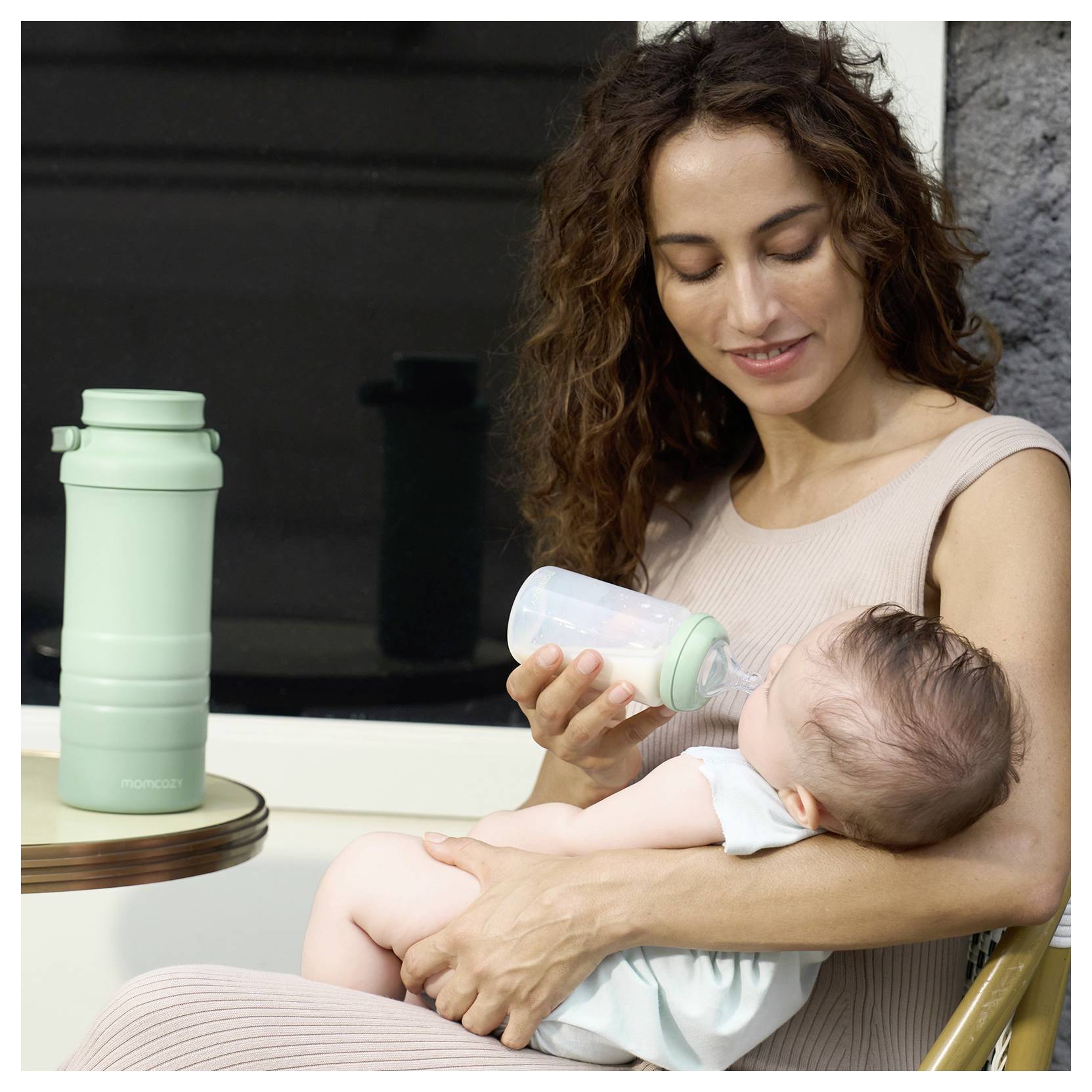 A woman in a beige dress feeds a baby with a bottle while sitting. A green water bottle is on a nearby table.