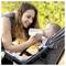 A woman gently feeds a baby with a bottle while the baby sits in a stroller. They are outdoors, surrounded by greenery.