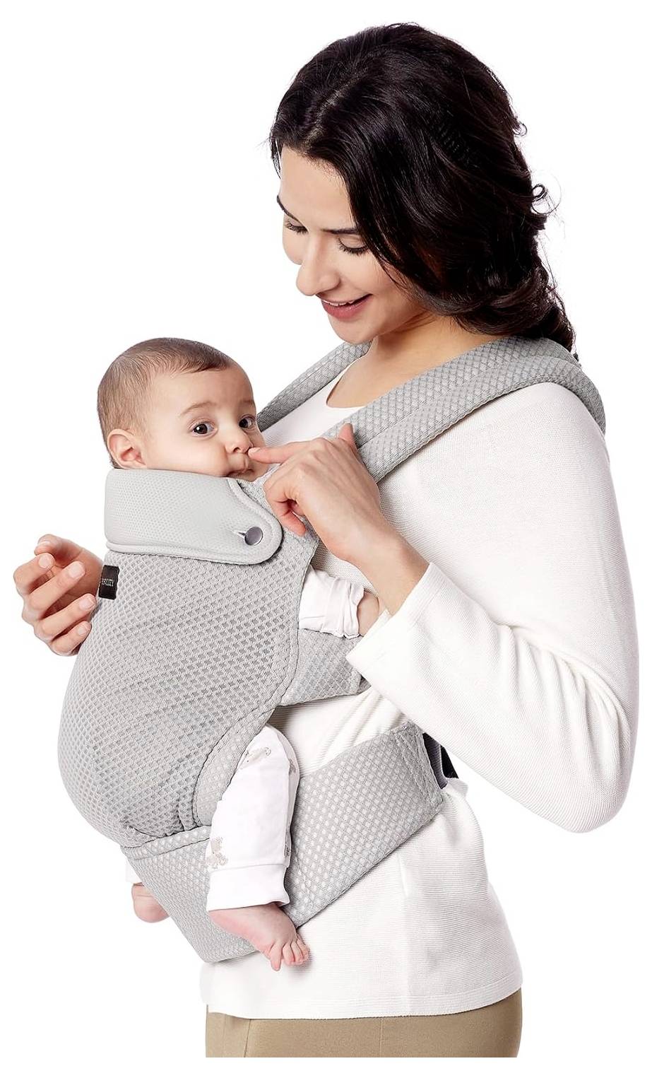 A woman smiles while holding an infant in a front baby carrier. The baby looks content and secure, facing outward as the woman supports them.
