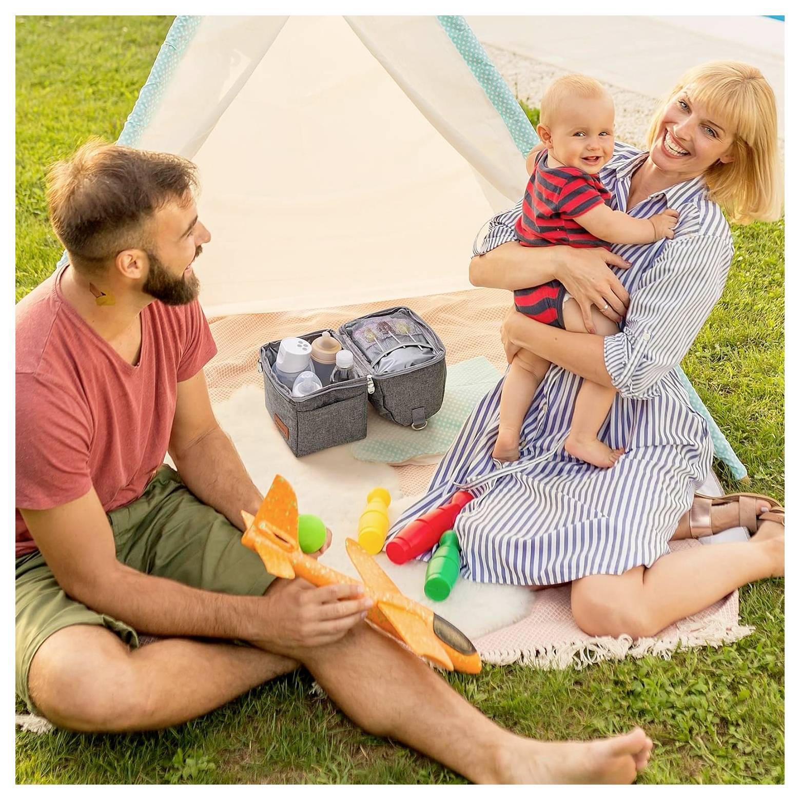 A family of three sits outside a small tent on grass. The mother holds a smiling baby while the father plays with toy tools. A picnic bag is nearby.