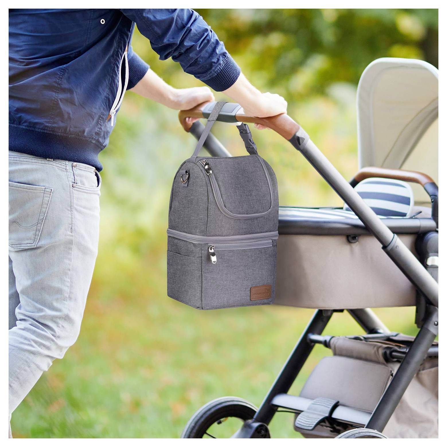 A person pushes a stroller in a park while carrying a grey insulated lunch bag attached to the stroller's handle.