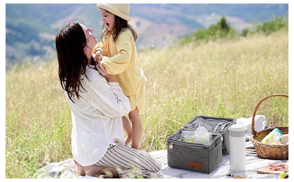 A woman and child enjoy a picnic outdoors. The woman holds the child who is wearing a yellow outfit and hat. A picnic basket and bag are nearby.