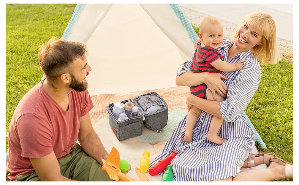A family having a picnic outdoors. A woman holds a smiling baby, while a man sits nearby with toys and a cooler bag on a blanket.