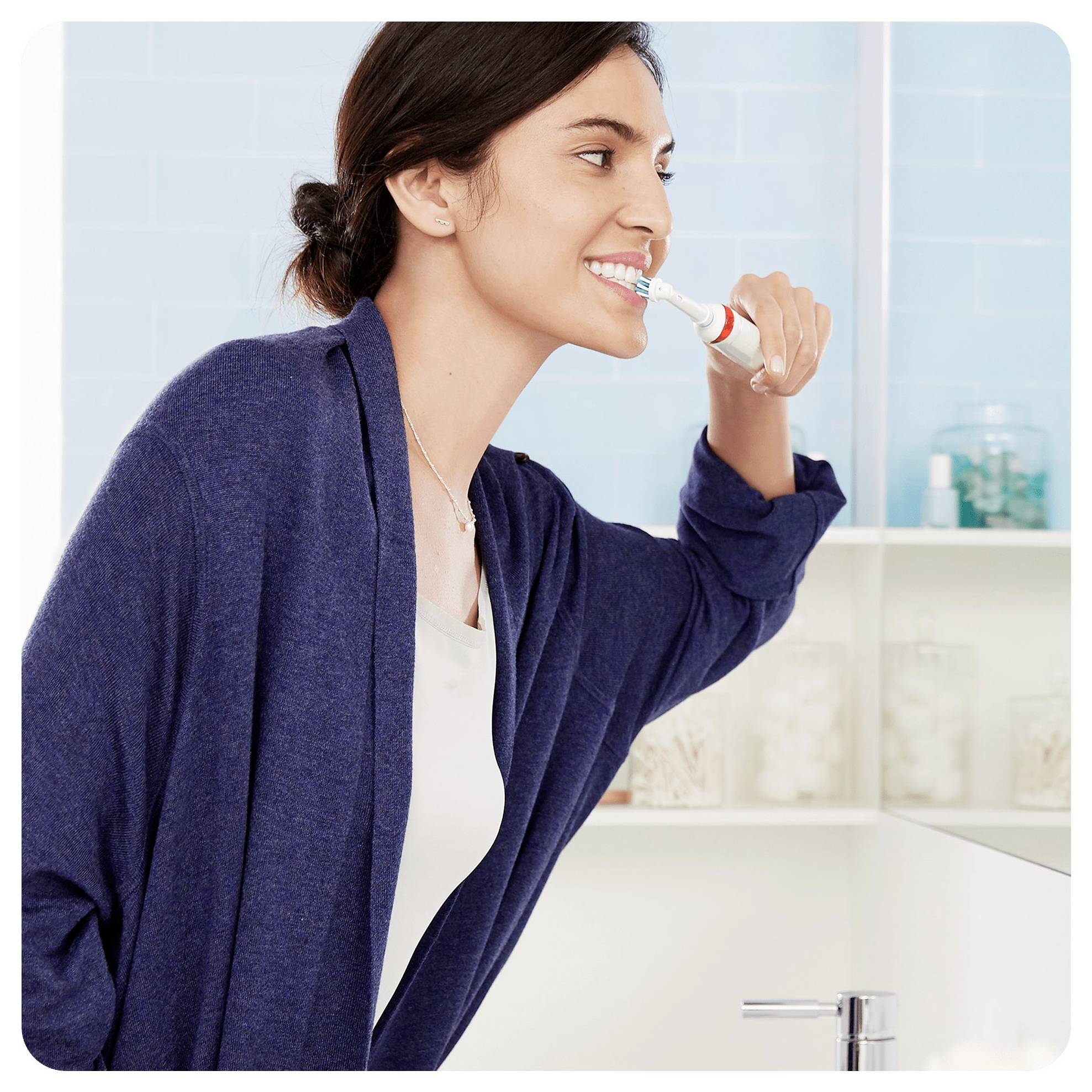 A person in a blue robe is brushing their teeth with an electric toothbrush in a bathroom setting, indicating a morning routine.