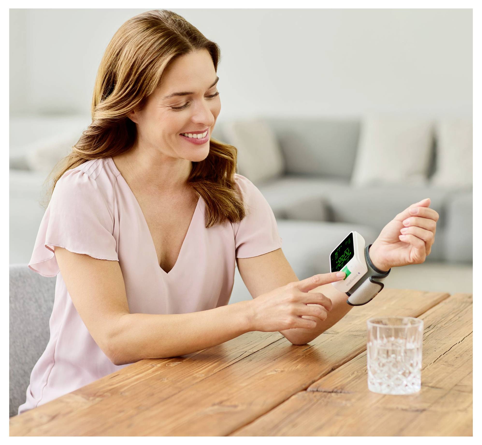 A woman sitting at a table, smiling while checking her blood pressure with a wrist monitor, next to a glass of water.