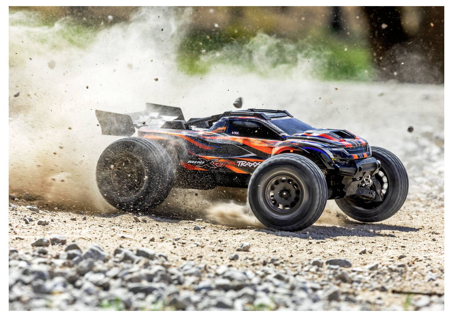 A remote-controlled car speeds across a gravel path, kicking up dust, with vibrant red and black markings on its body.