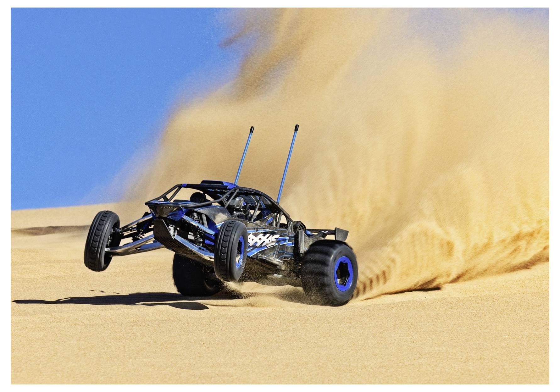 A remote-controlled car powers through a sandy dune, kicking up a large cloud of sand against a clear blue sky.