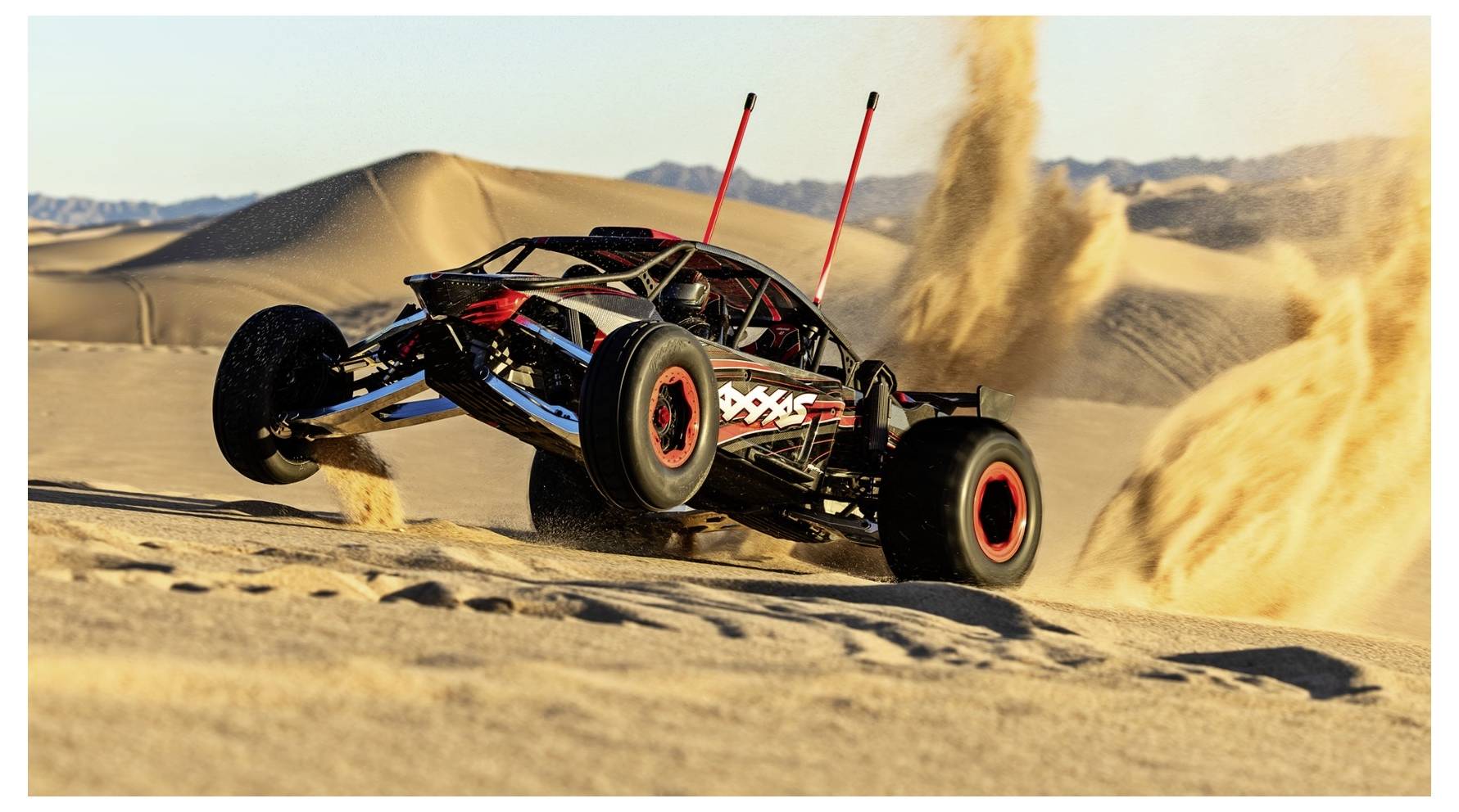 A remote-controlled car drives fast over sand dunes, kicking up sand behind it. The vehicle is black with red and white decals.