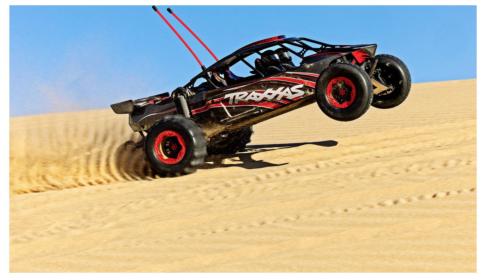 A dune buggy with red accents and 'TRAXXAS' branding leaps off a sandy dune under a clear blue sky.
