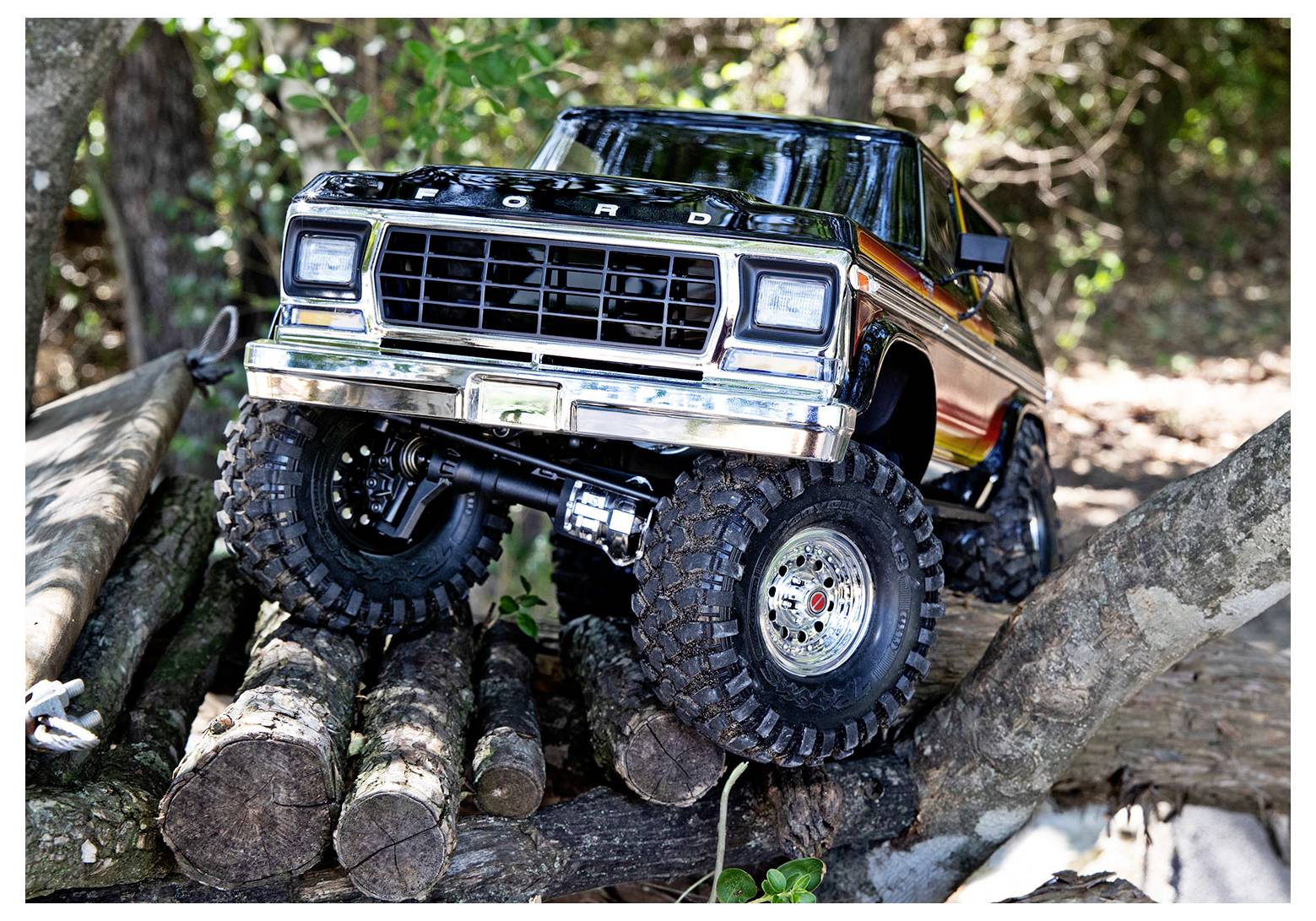 A toy truck with large, rugged tires navigates over stacked logs in a wooded area, showcasing its off-road capability.
