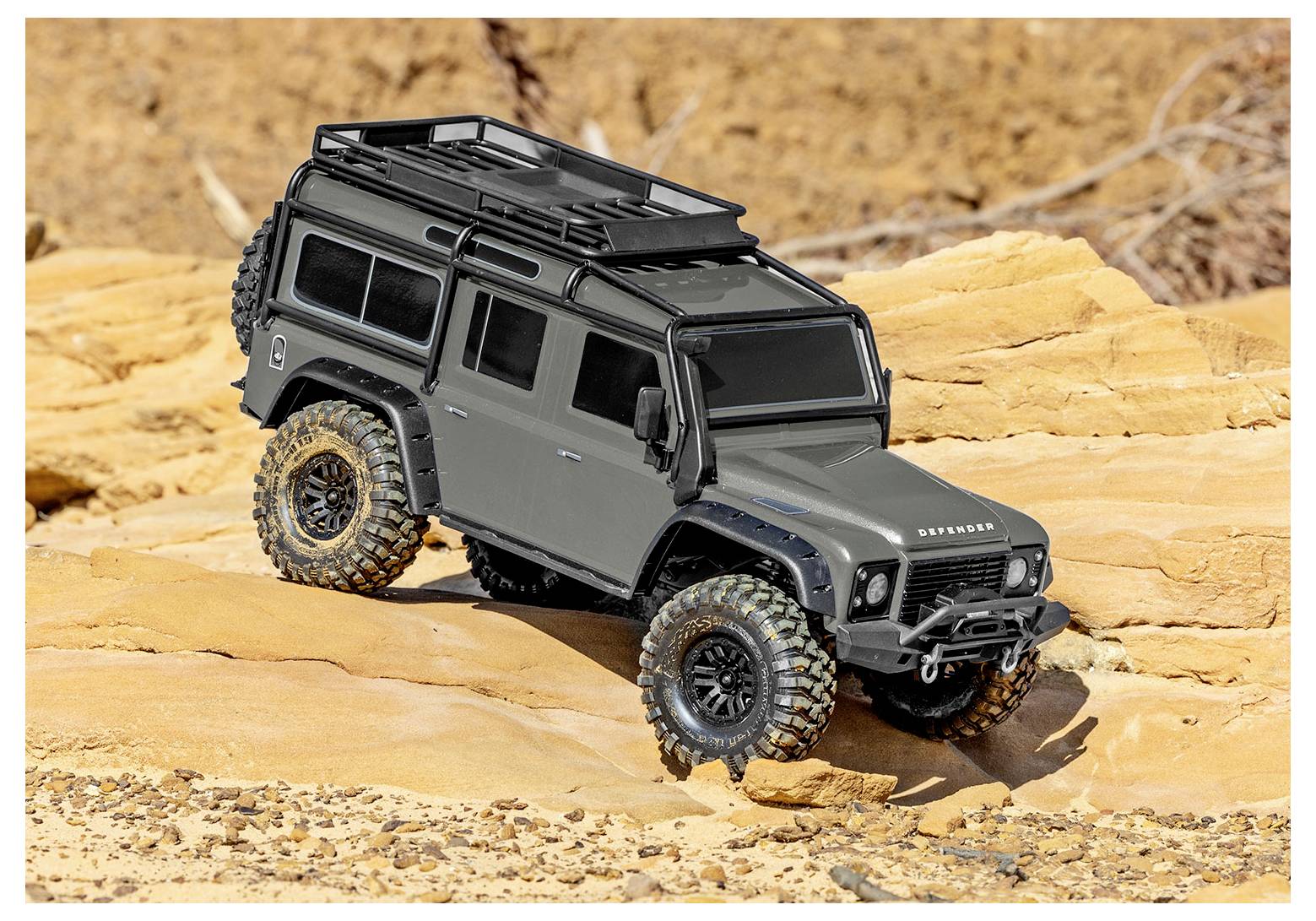 A grey off-road toy vehicle on rocky terrain with rugged tires and roof rack, showcasing its capability for navigating rough surfaces.