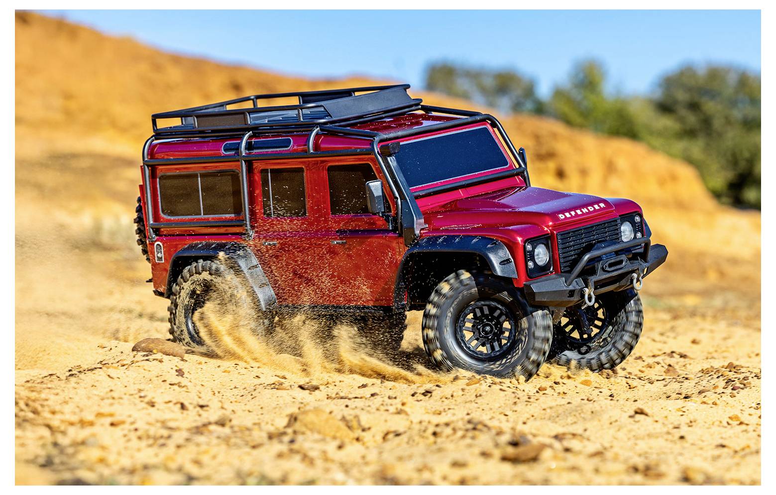 A red remote-controlled off-road vehicle drives through a sandy, rugged terrain, kicking up dust, with orange cliffs and a blue sky in the background.