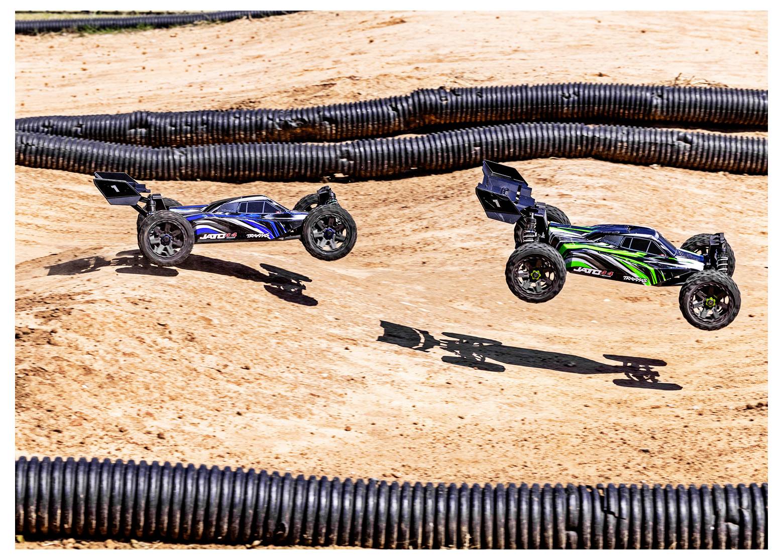 'Two remote-controlled cars, one blue and one green, jump over a dirt track in a race, casting shadows on the ground.'