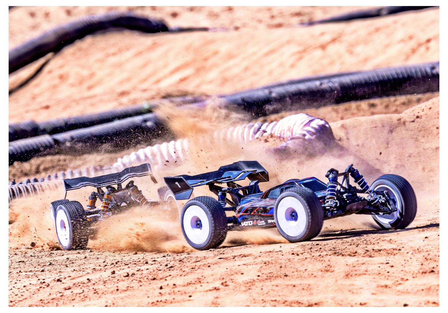 Two remote-controlled cars race on a dirt track, kicking up dust. The front car is blue and black, closely followed by a red car.